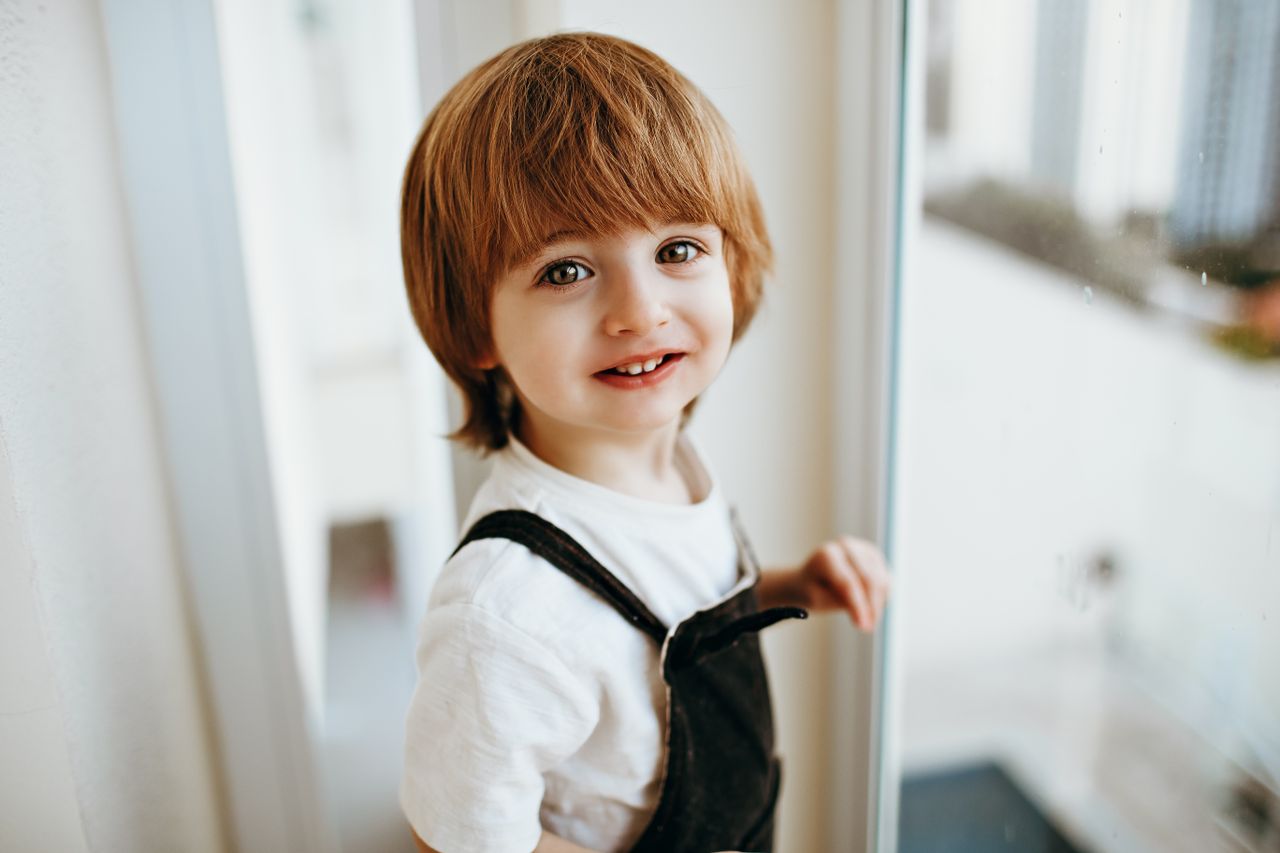 A joyful child in casual attire smiles while standing by a bright window indoors.