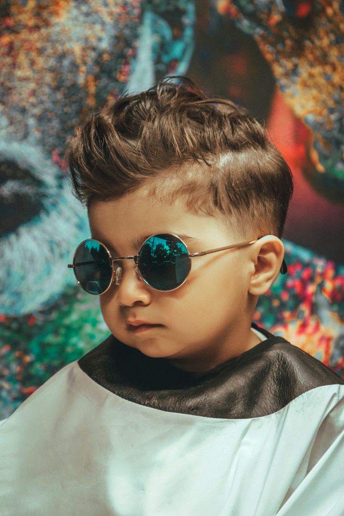 Young child with a trendy haircut and sunglasses, posing indoors against an artistic backdrop.