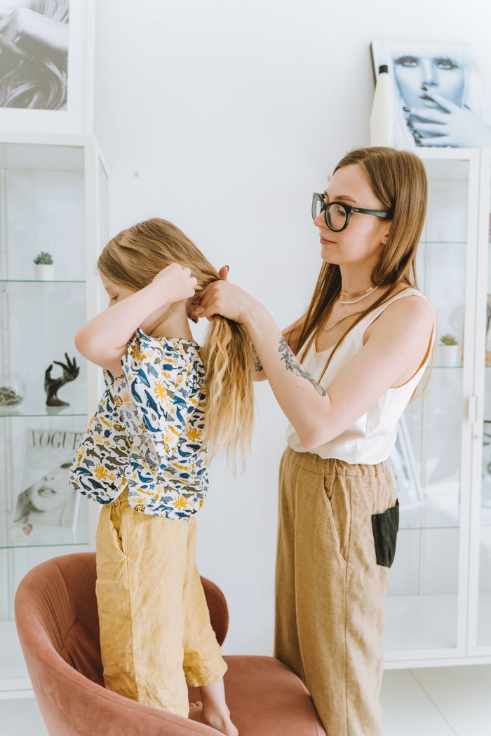 A mother braids her daughter's hair in a modern room with magazine decor. Indoors, warm atmosphere.